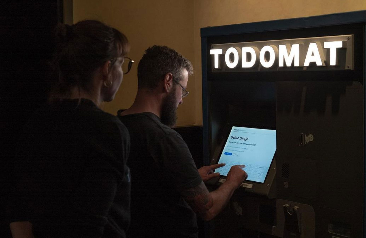 Two people are using the Todomat machine. One person is touching the touchscreen with two fingers, while the other person stands behind looking at the screen.