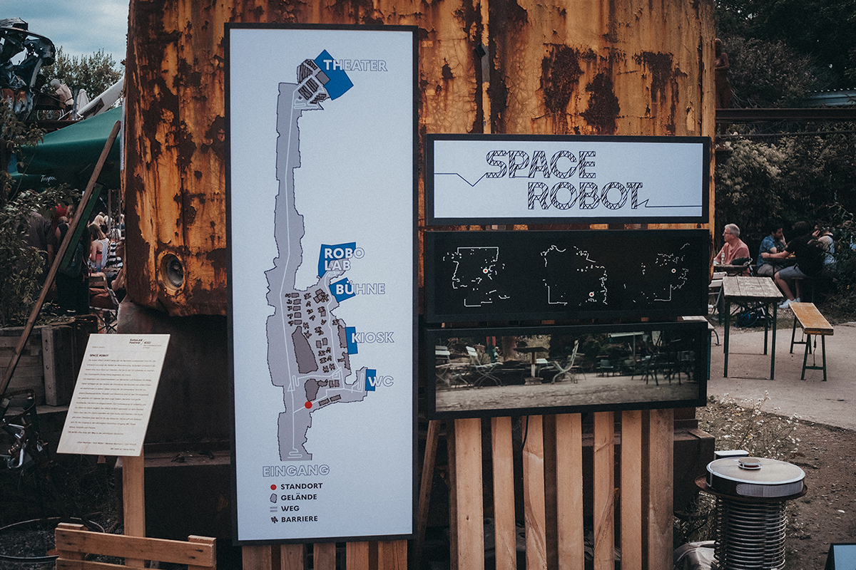 A large map displayed on a wooden structure at the Odonien festival. The map outlines the festival grounds, highlighting key areas such as the entrance, restrooms, kiosk, stage, Robolab, and theater. A white line traces the main path through the festival, indicating accessible routes. To the right, three screens showcase the process of how the map was created using a modified vacuum cleaner robot. The robot itself is also visible next to the screens.