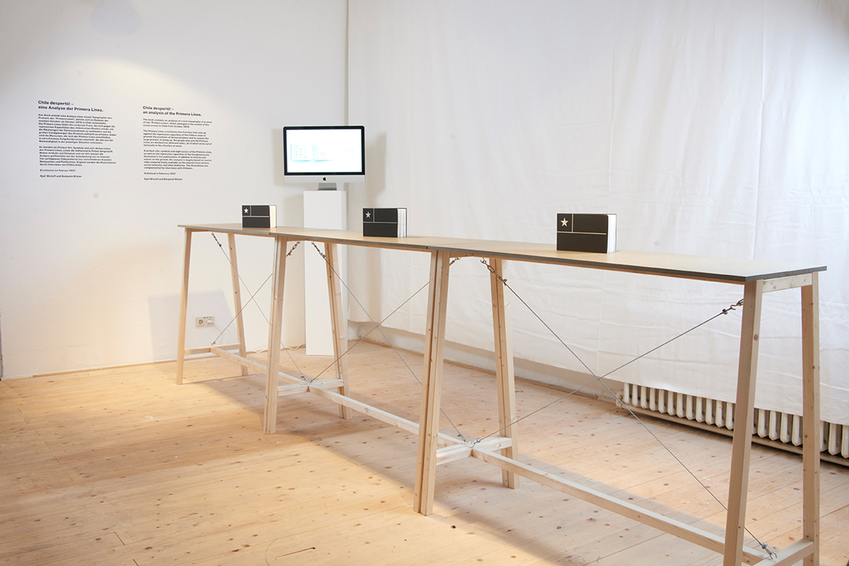 A long wooden table with four of the books arranged on it.  There is a computer monitor on a stand behind the main table. The room has white walls and a wooden floor.