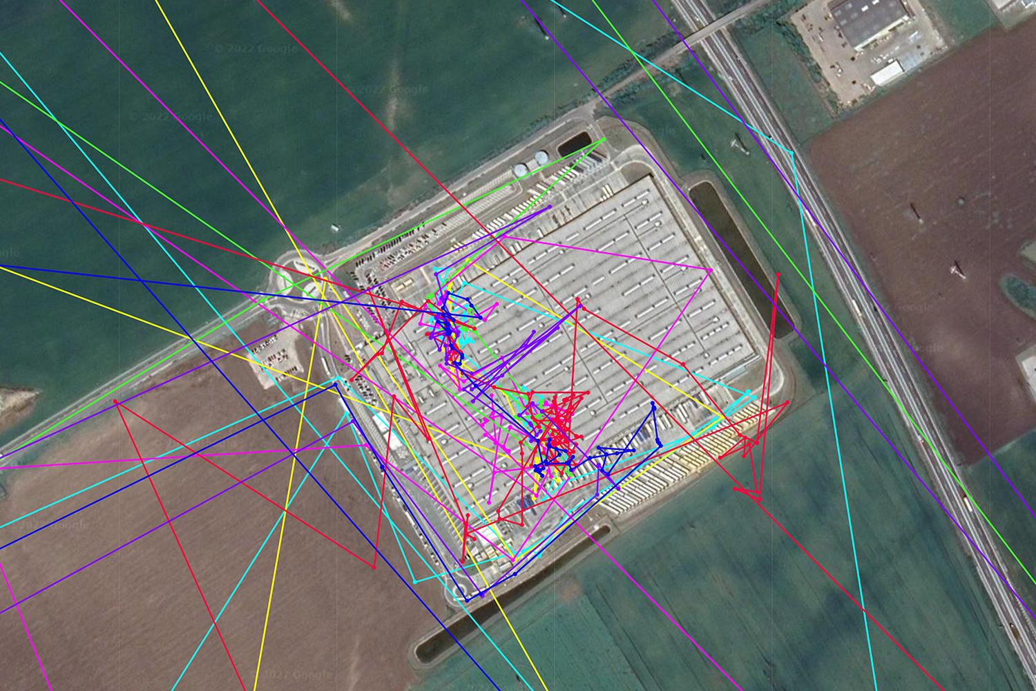 An aerial photograph of Amazon's BTS2 return warehouse in Bratislava. The warehouse is surrounded by fields and roads. Numerous colored lines converge on the warehouse from various directions. These lines represent the GPS-tracked journeys of returned packages to the warehouse.