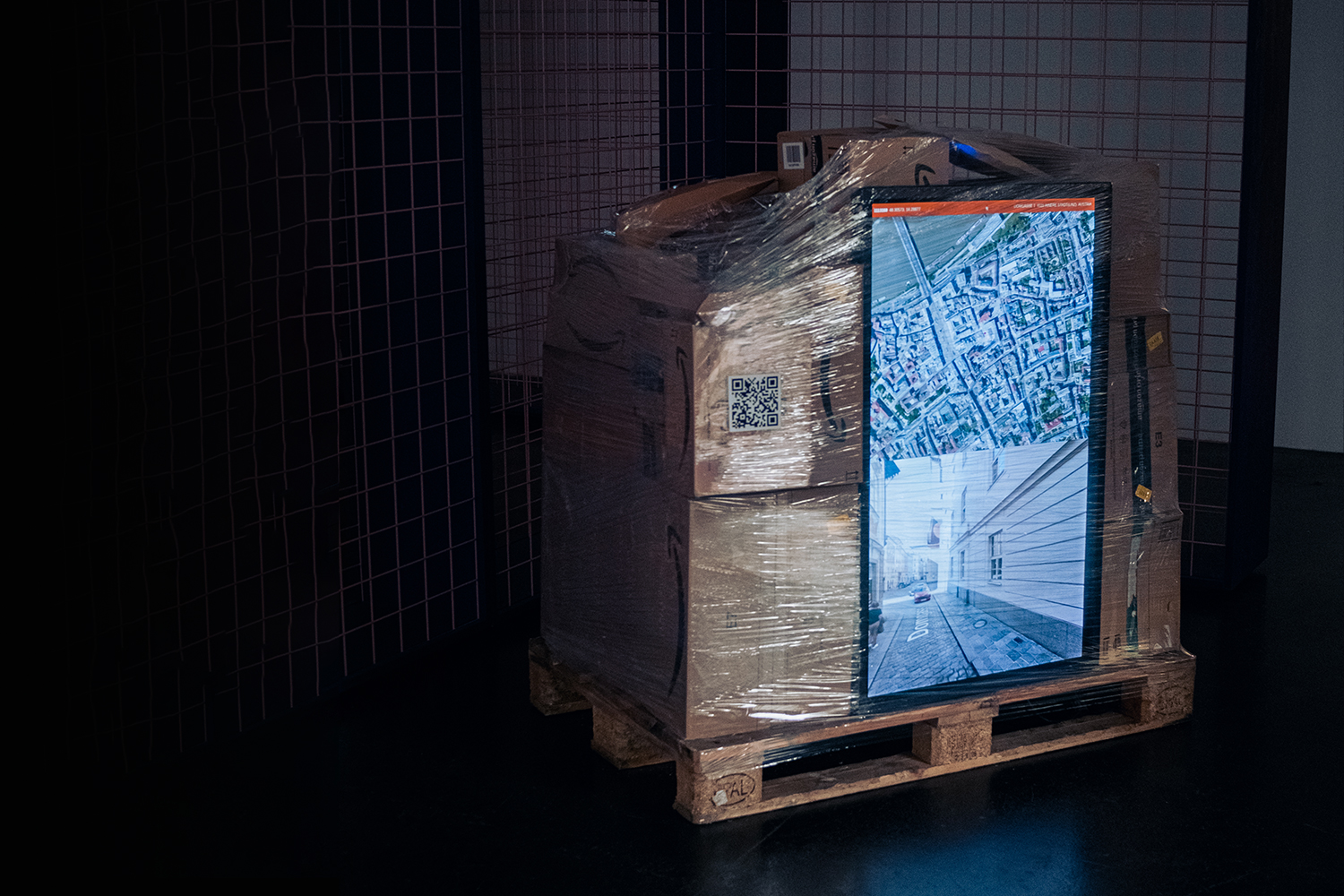A large, flat-screen monitor displaying a map and a street view is mounted on a pallet. The pallet is stacked with cardboard boxes wrapped in plastic.