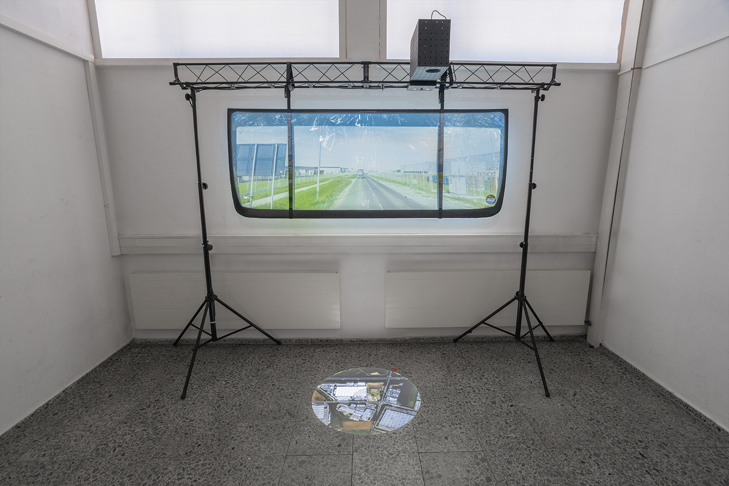 A large truck windshield is mounted on a wall in a white room. A projector above the windshield projects a moving image of a road onto the windshield, simulating a driving view. On the floor below, a circular projection displays live locations of returned trackers.