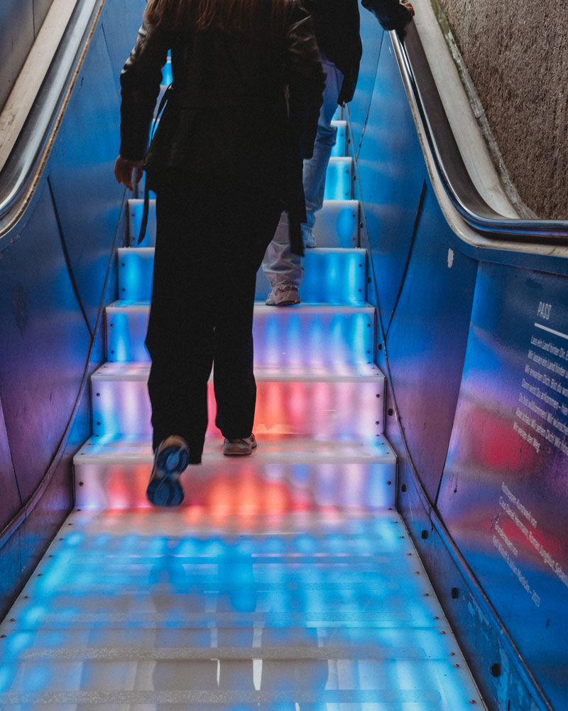 A person walking up the illuminated stairs. The stairs are brightly lit with a pattern of blue and red lights. The person is wearing dark clothing.