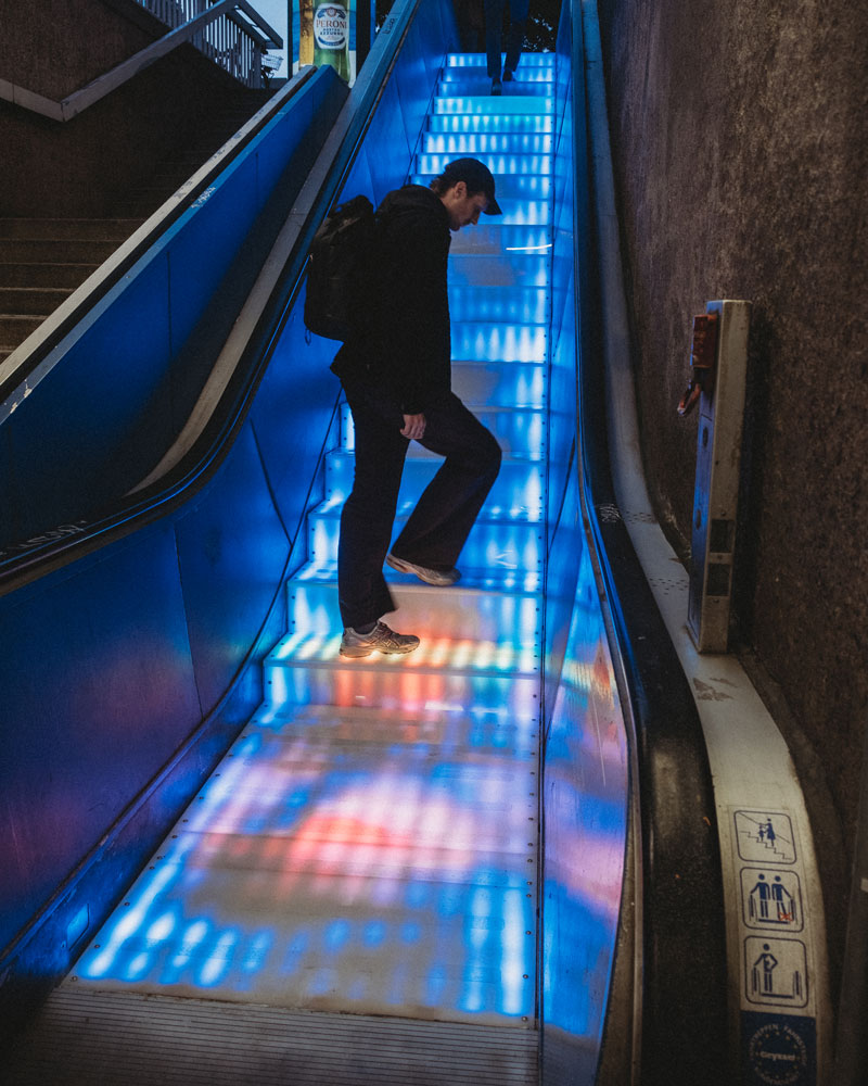 Another person walking up the illuminated stairs. The stairs are also brightly lit with a pattern of blue and red lights. The person is also wearing dark clothing.