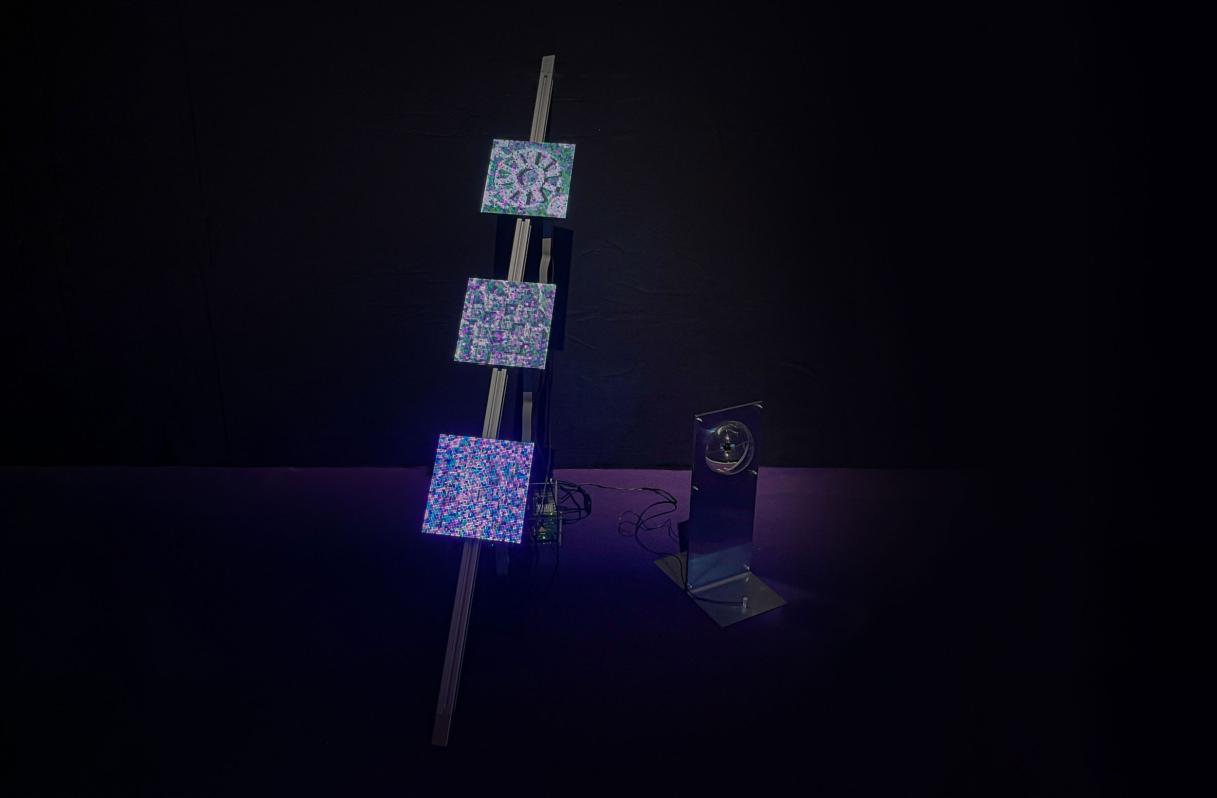 Art installation featuring three illuminated square panels mounted on a tilted metallic structure, emitting colorful pixelated satellite imagery. Nearby, a standalone reflective device sits on a small stand, all set against a dark background.